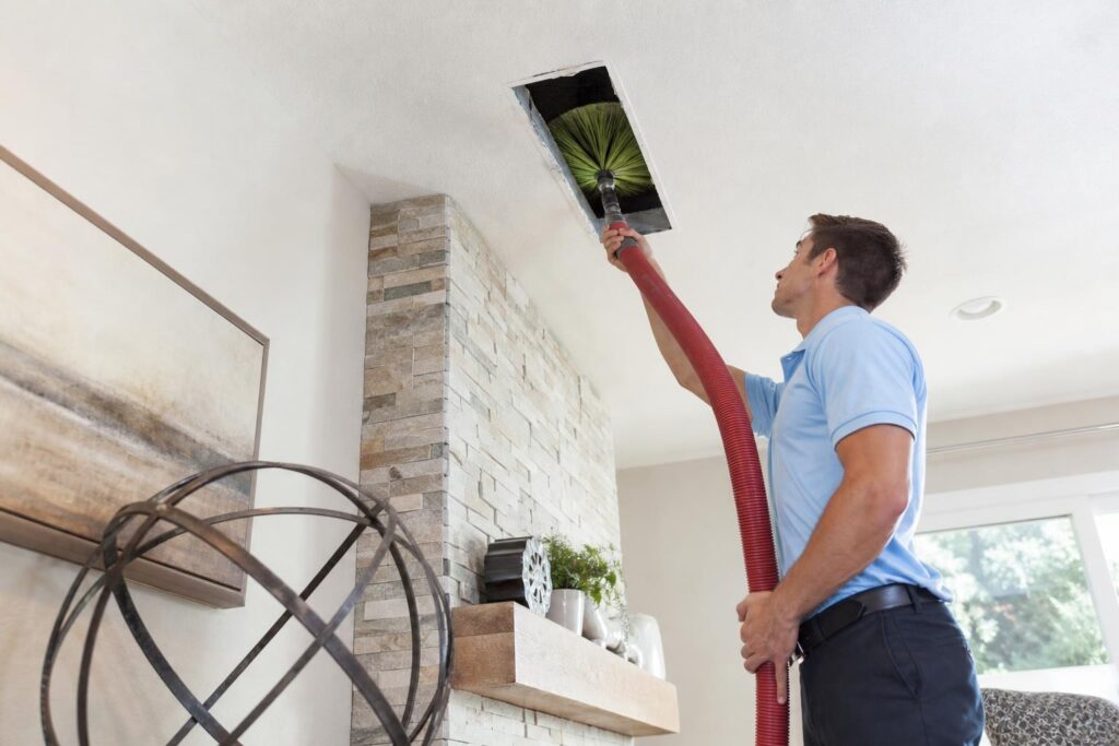 Service technician in a blue polo shirt using a long red vacuum hose and green rotating brush to clean a ceiling air duct in a modern home.